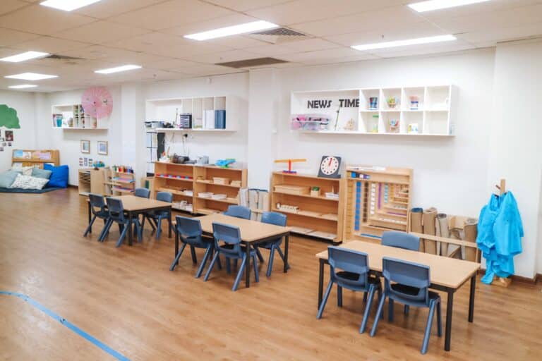 Montessori Academy Alexandria childcare classroom with three child-sized tables, four chairs at each, and Montessori shelves in the background.